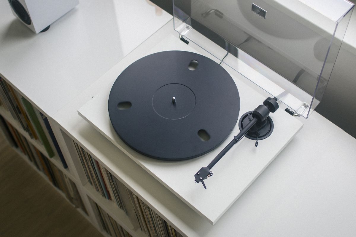 Ein weißer Plattenspieler mit schwarzem Plattenteller steht auf einem Regal voller Schallplatten. Der Tonarm ist seitlich abgelegt, die transparente Staubschutzhaube ist geöffnet.
