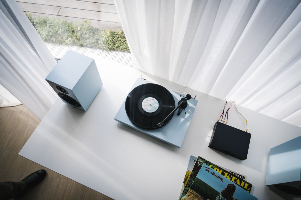 Ein hellblauer Plattenspieler mit aufgelegter Schallplatte steht auf einem weißen Tisch, flankiert von zwei passenden Lautsprechern. Daneben liegt ein Stapel Vinylhüllen, im Hintergrund fällt weiches Tageslicht durch weiße Vorhänge.