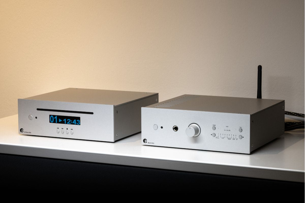 Zwei silberne HiFi-Komponenten auf einem Sideboard: links ein CD-Player mit blauem Display, rechts ein Verstärker/Receiver mit Lautstärkeregler und Antenne vor einer beigen Wand.