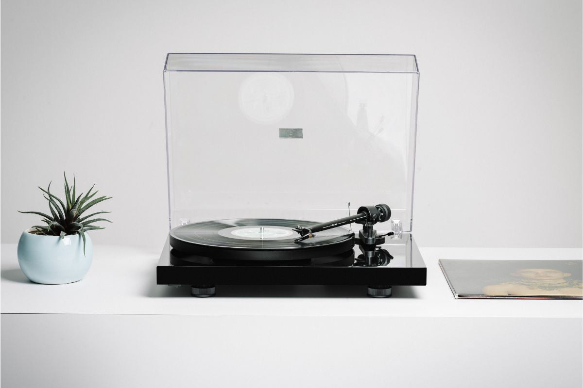 Moderner Schallplattenspieler mit transparenter Abdeckung und aufgelegter Vinyl-Schallplatte auf weißem Sideboard, daneben eine kleine Pflanze und ein Plattencover