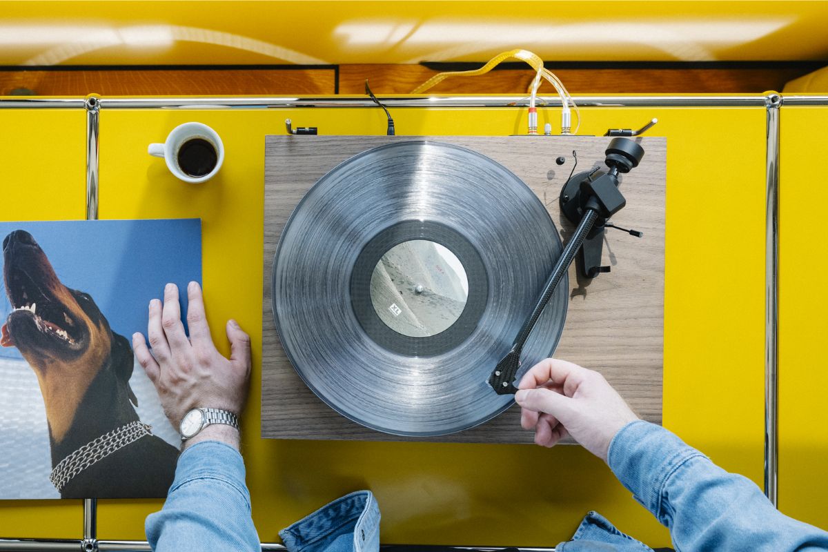 High-End-Plattenspieler auf gelber Ablagefläche, von oben fotografiert, während eine Person den Tonarm auf eine transparente Vinyl-Schallplatte setzt