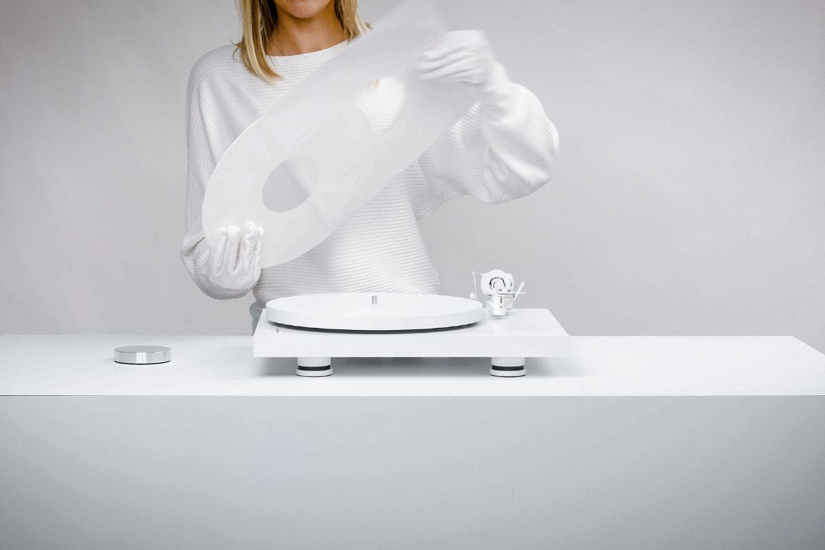 Eine Person in weißer Kleidung und weißen Handschuhen legt eine transparente Schallplatte auf einen weißen Plattenspieler in minimalistischer Umgebung.
