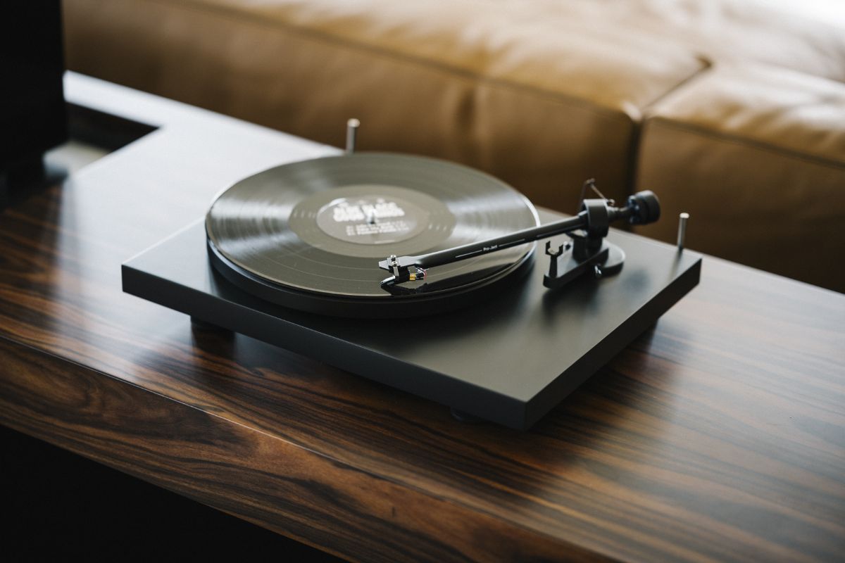 Ein schwarzer Plattenspieler mit aufgelegter Vinyl-Schallplatte steht auf einem edlen Holztisch. Der Tonarm ist abgesenkt, im Hintergrund ist eine braune Ledercouch unscharf zu sehen.