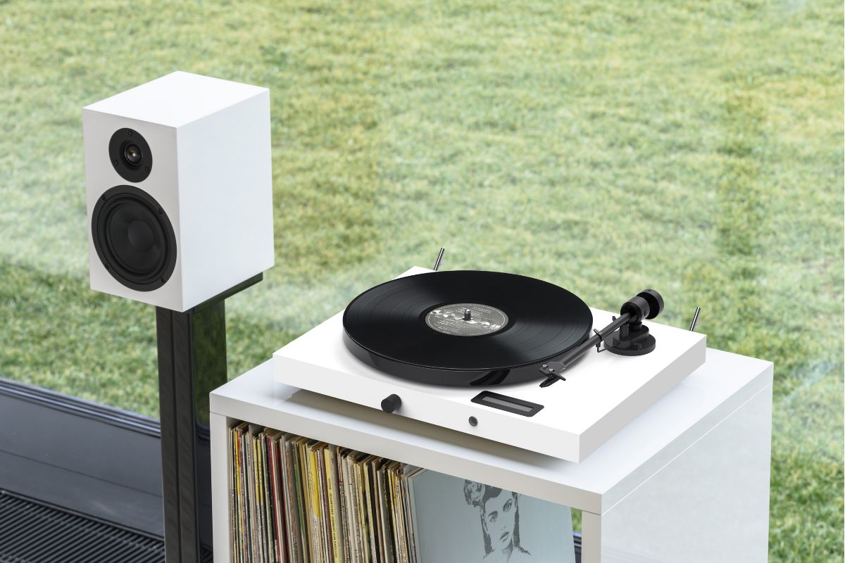 Weißer Plattenspieler mit schwarzer Schallplatte auf einem Regal mit Schallplatten, daneben ein weißer Lautsprecher vor einer Glasfront mit Blick ins Grüne.