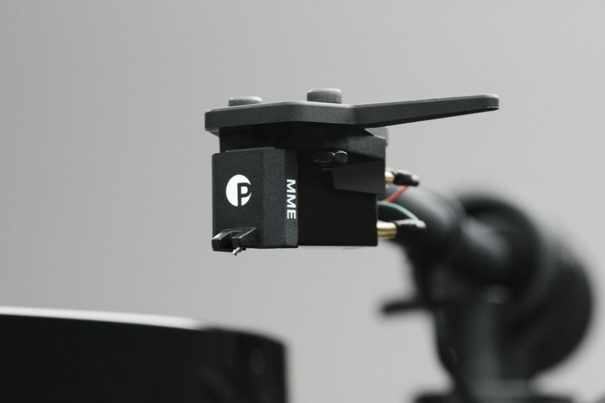 Nahaufnahme eines schwarzen Tonabnehmers mit der Aufschrift „MME“ und dem Pro-Ject-Logo, montiert am Tonarm eines Plattenspielers vor neutralem grauem Hintergrund.