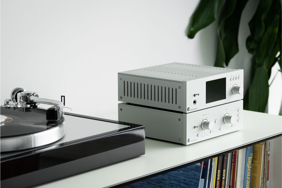 Ein moderner Plattenspieler steht links im Bild, daneben zwei silberne HiFi-Komponenten mit Drehreglern und Display auf einem weißen Regal. Darunter sind Schallplatten zu sehen, im Hintergrund steht eine grüne Zimmerpflanze.