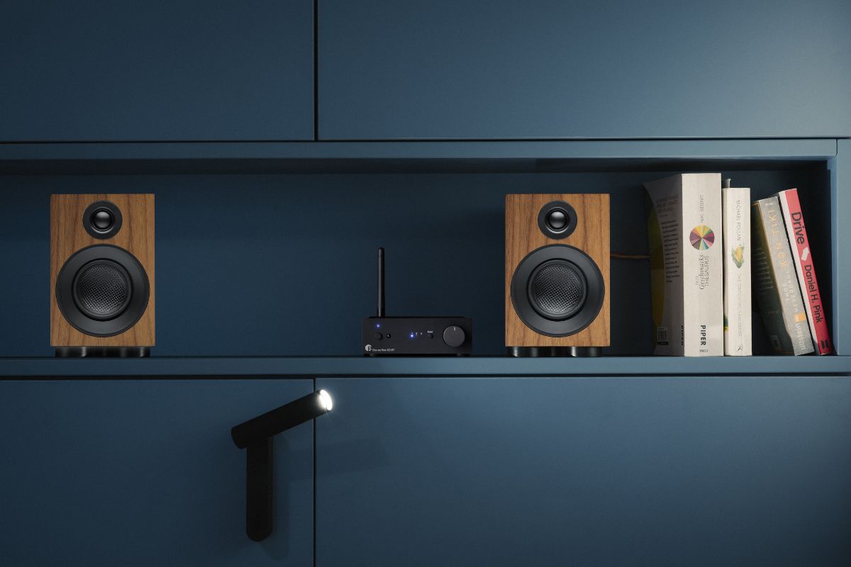 Modernes HiFi-Setup in einem blauen Regal mit zwei kompakten Lautsprechern in Holzoptik und einem zentralen Verstärker, umgeben von Büchern