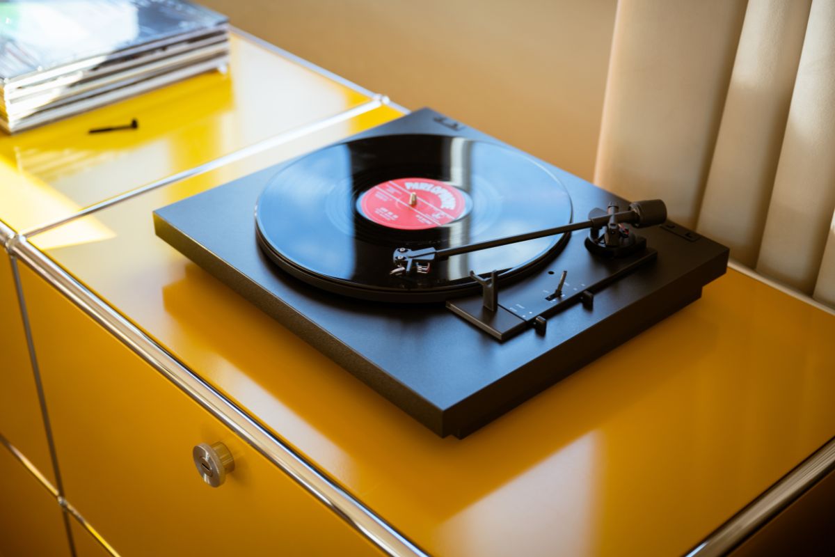 Schwarzer Plattenspieler mit aufgelegter Schallplatte auf einem gelben Sideboard neben einem Stapel weiterer Vinylplatten.