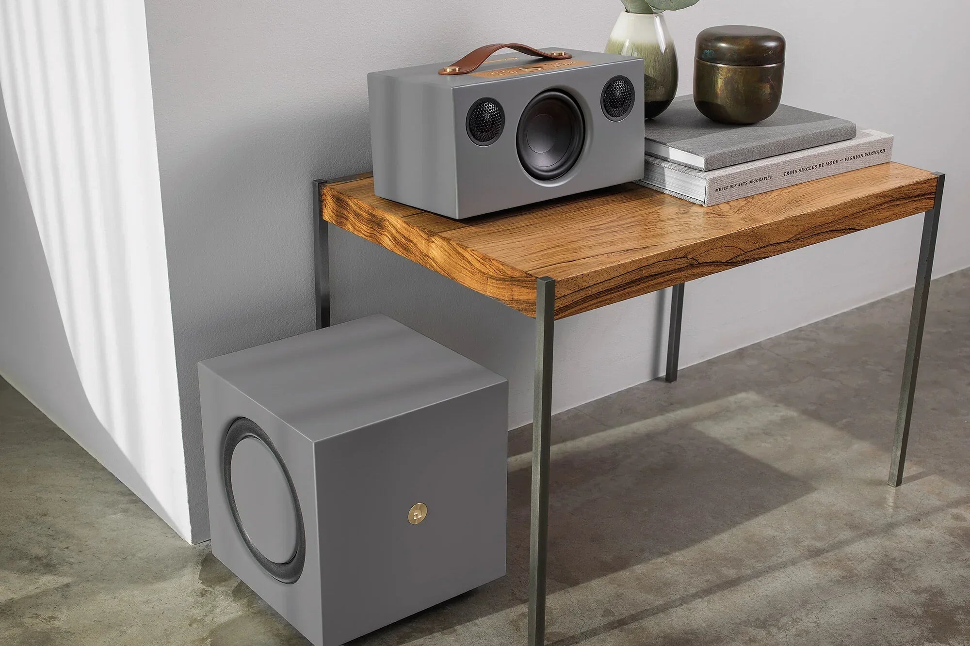 Grauer Smart-Home-Lautsprecher mit Subwoofer auf einem Holzregal im Wohnzimmer