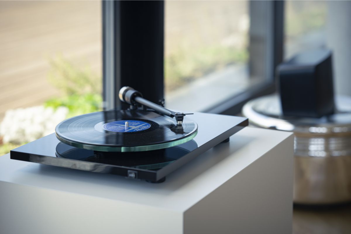 Moderner HiFi-Plattenspieler mit Glassubteller und aufgelegter schwarzer Vinyl-Schallplatte, platziert auf einem weißen Sideboard vor einem großen Fenster.