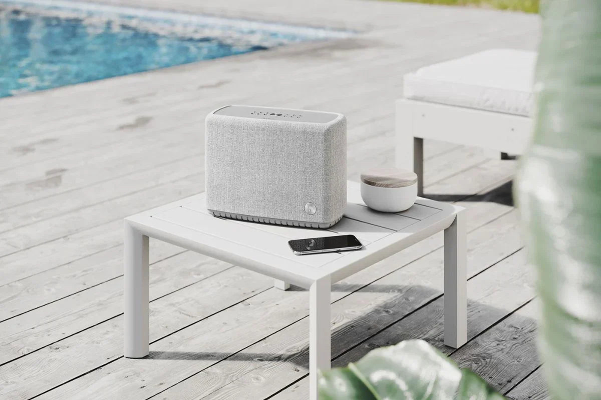 Tragbarer Multiroom-Lautsprecher in Hellgrau von Audio Pro auf einem weißen Terrassentisch am Pool